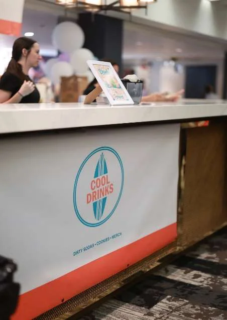 The Cool Drinks catering booth at an indoor event, with the logo on the front of the counter and a team member serving behind it
