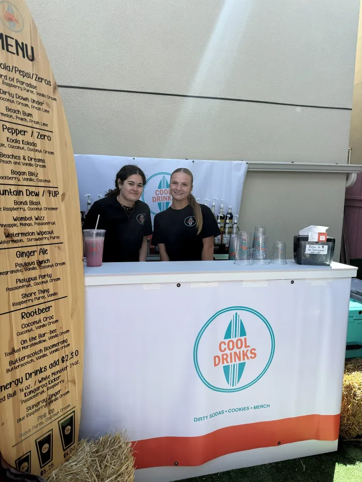 Two team members behind the Cool Drinks booth next to a surfboard-shaped drink menu