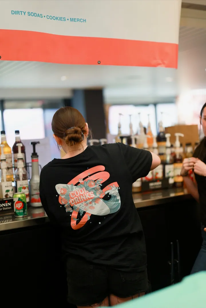 The back of a Cool Drinks team shirt, printed with a surfboard, flowers and a wave, as a team member mixes drinks